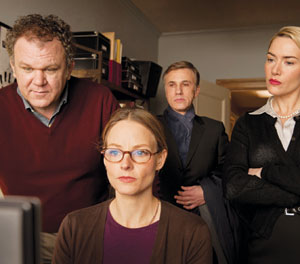 Four adults stand closely together, looking intently at a computer screen. One woman with glasses sits in front, while two men and another woman stand behind her, all appearing serious and focused.