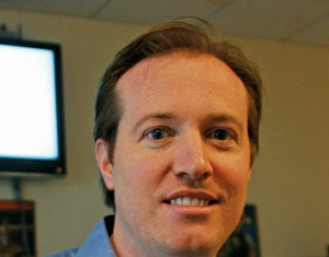 A man with light brown hair wearing a light blue collared shirt smiles at the camera indoors, with a television screen and office background visible behind him.