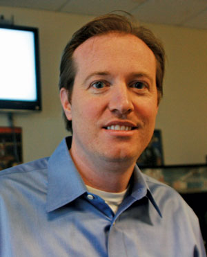 A man with light brown hair wearing a light blue collared shirt smiles at the camera indoors, with a television screen and office background visible behind him.