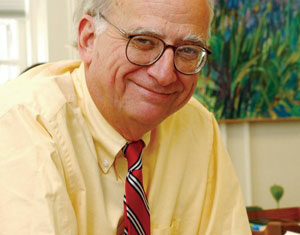An older man with glasses, wearing a yellow shirt and red striped tie, sits at a desk with papers and a keyboard, smiling at the camera.