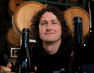 A person with curly hair smiles while holding a wine bottle. Other wine bottles are visible in the foreground, with wooden barrels stacked in the background.