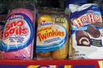 Packages of Hostess snack cakes on a store shelf, including Sno Balls, Golden Twinkies, and HoHos. Each package displays the product and colorful branding.
