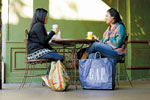 Two women sit at a small round table outdoors, drinking coffee and talking. Shopping bags, including a large blue GAP bag, are placed near their chairs. The background features a green wall.