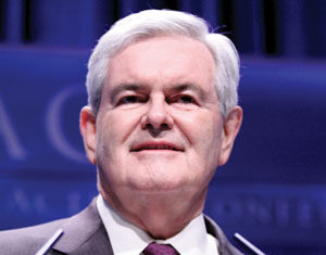 A man with short white hair in a dark suit, white shirt, and red tie stands at a podium, smiling slightly, with a blue background behind him.