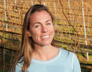 A woman with long, light brown hair and a light blue sweater smiles while standing outdoors in a vineyard with rows of grapevines in the background.