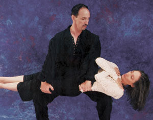 A man in black holds a woman in a white dress horizontally across his legs, as if performing a dance or illusion, against a purple studio backdrop.