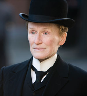 A person wearing a black bowler hat, white shirt with a high collar, black jacket, and black tie looks into the distance with a neutral expression. The background is softly blurred.