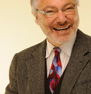 A smiling older man with gray hair and a beard, wearing glasses, a gray tweed jacket, a colorful patterned tie, and a white shirt against a light background.