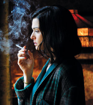 A woman with dark hair in a plaid coat stands indoors, holding a lit cigarette near her face as smoke curls upward. Warm, soft lighting and a vintage lamp create a moody, contemplative atmosphere.