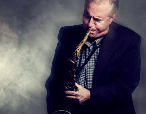 Older man wearing a dark blazer and checkered shirt playing a saxophone with a focused expression against a smoky, dimly lit background.