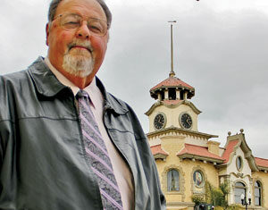An older man wearing glasses, a leather jacket, shirt, and patterned tie stands in front of a historic building with a clock tower and ornate architectural features on a cloudy day.
