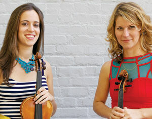 Two women standing in front of a light gray brick wall, each holding a violin. One wears a striped dress with a yellow belt and blue necklace; the other wears a red dress with turquoise accents. Both are smiling at the camera.