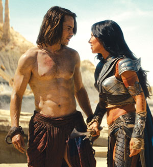 A shirtless man with long hair stands facing a woman in elaborate armor in a desert setting, looking intensely at each other. Rocky formations are visible in the background.