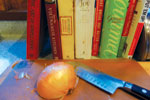 A whole onion and a chef’s knife rest on a cutting board in front of a row of colorful cookbooks on a kitchen counter.
