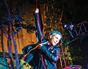 A singer in a black leather jacket passionately performs on stage, holding a red microphone cable, while a drummer plays in the background. The stage is outdoors at night, with colorful lighting and an enthusiastic crowd.