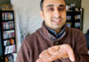 A person in a brown sweater smiles and holds a small object in their open palm, standing in a living room with shelves of books and DVDs in the background.