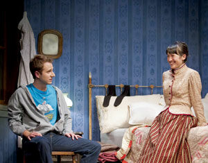 A young man in casual clothes sits on a chair next to a bed, while a woman in a vintage striped dress sits on the bed, smiling. The room has blue patterned wallpaper and a small mirror on the wall.