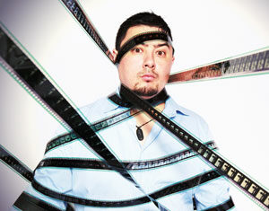 A man wearing a light blue shirt is wrapped in strips of photographic film that cover his torso, arms, and head. He looks directly at the camera with a surprised expression against a plain light background.