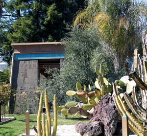 A small building with ancient Egyptian-style hieroglyphics on its front stands behind green trees, cacti, and desert plants in a sunlit garden.