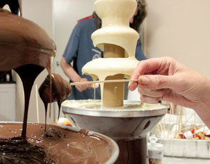 A person dips a fruit skewer into a bowl of melted chocolate near a chocolate fountain, with a white chocolate fountain and another person visible in the background.