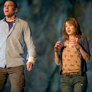 A man and a woman stand side by side looking alarmed. The man has a bandaged wrist and both appear tense, with the woman holding her hands up in fear. The background is blurry and dark.