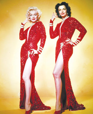 Two women stand side by side against a yellow background, wearing matching red sequined dresses with thigh-high slits and red high heels. They both pose with one hand raised and the other on their hip, exuding glamour.