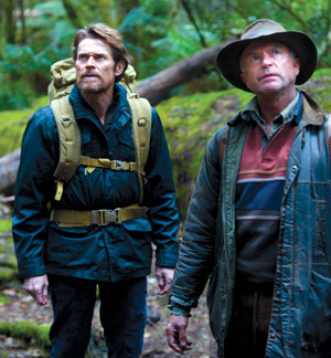 Two men stand in a lush, green forest; one wears a backpack and dark jacket, while the other wears a hat and layered clothing. Both are looking upward with serious expressions.