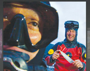 Two people in diving gear; one is close to the camera with a mask on, while the other, further back and wearing a red drysuit, smiles and holds a large knife outdoors.