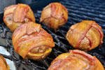 Six bacon-wrapped meatballs are cooking on a grill, with crispy strips of bacon wrapped around each ball, creating a golden-brown and appetizing appearance.