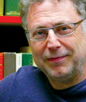 A middle-aged man with gray curly hair, glasses, and a slight smile, wearing a blue shirt, is seen in front of a bookshelf filled with various colorful books.