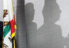 A California state flag next to a curtain showing the shadows of two people in profile, suggesting a behind-the-scenes or private conversation.