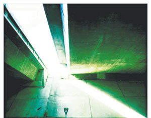 A view under a highway overpass with concrete walls and beams. Sunlight streams in from the left, casting bright and dark shadows across the scene. A parking meter stands by the empty road in the foreground.
