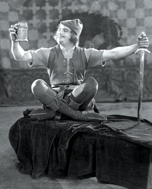 A man in medieval costume sits cross-legged on a platform, holding a large mug in one hand and a sword in the other, smiling and looking at the mug. The background features a patterned tapestry.