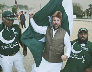 Three men pose together outdoors, two wearing dark green clothing with white crescent moon and star designs, one holding a large Pakistan flag and wearing a brown cap and vest. All appear to celebrate enthusiastically.