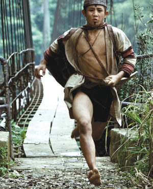 A young man wearing a tan robe and headband runs barefoot across a narrow, rustic bridge in a forested area, with a determined expression on his face.