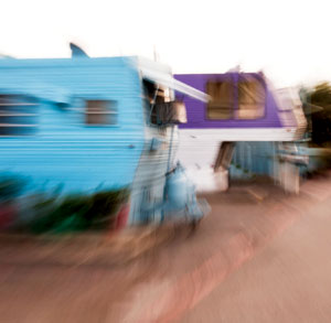 Blurred image of colorful trailers, primarily blue and purple, situated along a street or lot, with visible distortion suggesting motion or camera shake.