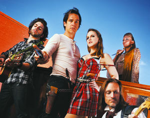 Five people pose outdoors in colorful outfits; two play guitars, one wears a western-style costume with a toy gun, a woman in a plaid dress stands confidently, and another person stands in the background, all against a blue sky.