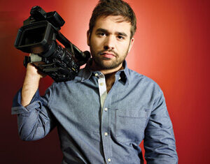 A man in a blue button-down shirt and dark jeans sits on a stool against a red wall, holding a large video camera on his shoulder and looking at the camera with a neutral expression.