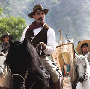 A man wearing a brown vest, white shirt, and wide-brimmed hat rides a black horse in a procession with others on horseback, carrying a religious banner featuring an image of the Virgin of Guadalupe.