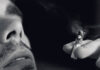 Close-up of a man’s face with stubble, holding a lit cigarette or joint near his mouth. Smoke is visible rising from the tip, and the image is in black and white.