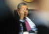 A man in a suit and red tie sits at a table, resting his head on one hand and partially covering his face. A water bottle and soda can are on the table in front of him.