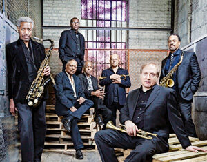 Seven men in suits pose with saxophones and trumpets on wooden pallets in an industrial, warehouse-like space with brick walls and a barred window in the background.