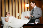 A woman lies in a bed covered by a white blanket, looking at a man in a suit who is standing beside her, gesturing with his hands in a room with striped wallpaper and a lit wall lamp.