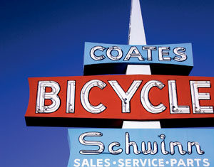 A retro vertical sign with COATES BICYCLE Schwinn SALES • SERVICE • PARTS against a blue sky, with palm trees and buildings visible in the background.