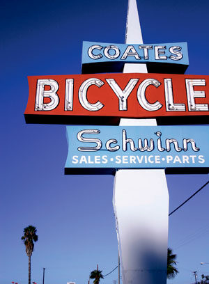 A retro vertical sign with COATES BICYCLE Schwinn SALES • SERVICE • PARTS against a blue sky, with palm trees and buildings visible in the background.