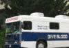 A blue and white blood donation bus labeled BLOODMOBILE and GIVE BLOOD is parked on a street, with trees and a building in the background.