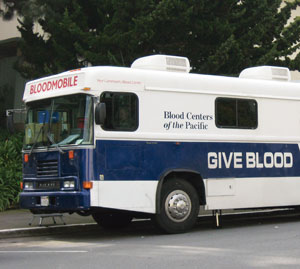 A blue and white blood donation bus labeled BLOODMOBILE and GIVE BLOOD is parked on a street, with trees and a building in the background.