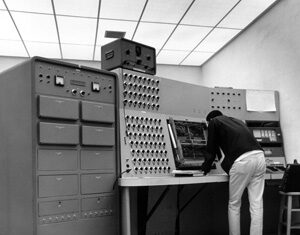 A person in white pants operates a large, vintage computer system with numerous dials, switches, and panels in a room with a tiled ceiling.
