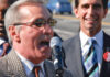 A man wearing sunglasses and a checkered jacket speaks into a microphone at an outdoor event, while another man in a suit and striped tie smiles in the background. Both wear red buttons that read No on 8.