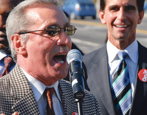 A man wearing sunglasses and a checkered jacket speaks into a microphone at an outdoor event, while another man in a suit and striped tie smiles in the background. Both wear red buttons that read No on 8.